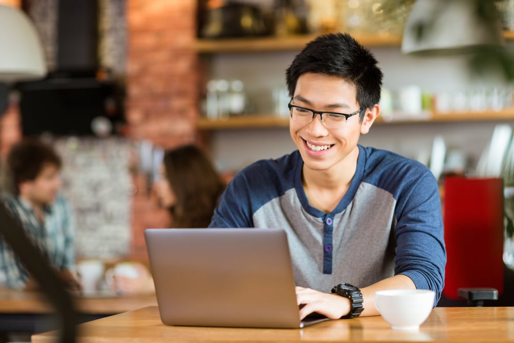 Happy cheerful young asian male in glasses smiling and using laptop in cafe-3