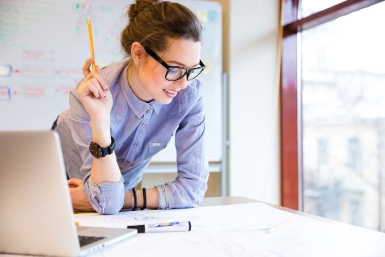 Happy young woman in glasses standing near the window in office and working with blueprint